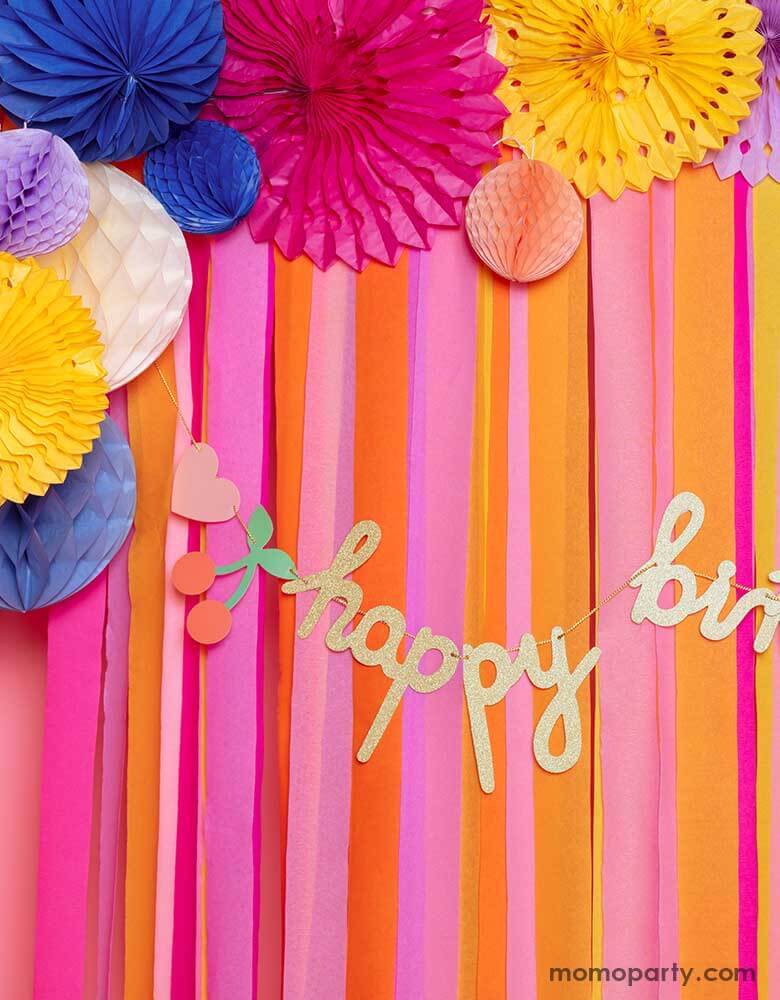 A closeup of a pink room decorated with colorful paper fans and streamers adorn with happy birthday banner with rainbows and cherries. A cheerful and festive decorations for birthdays of any age.