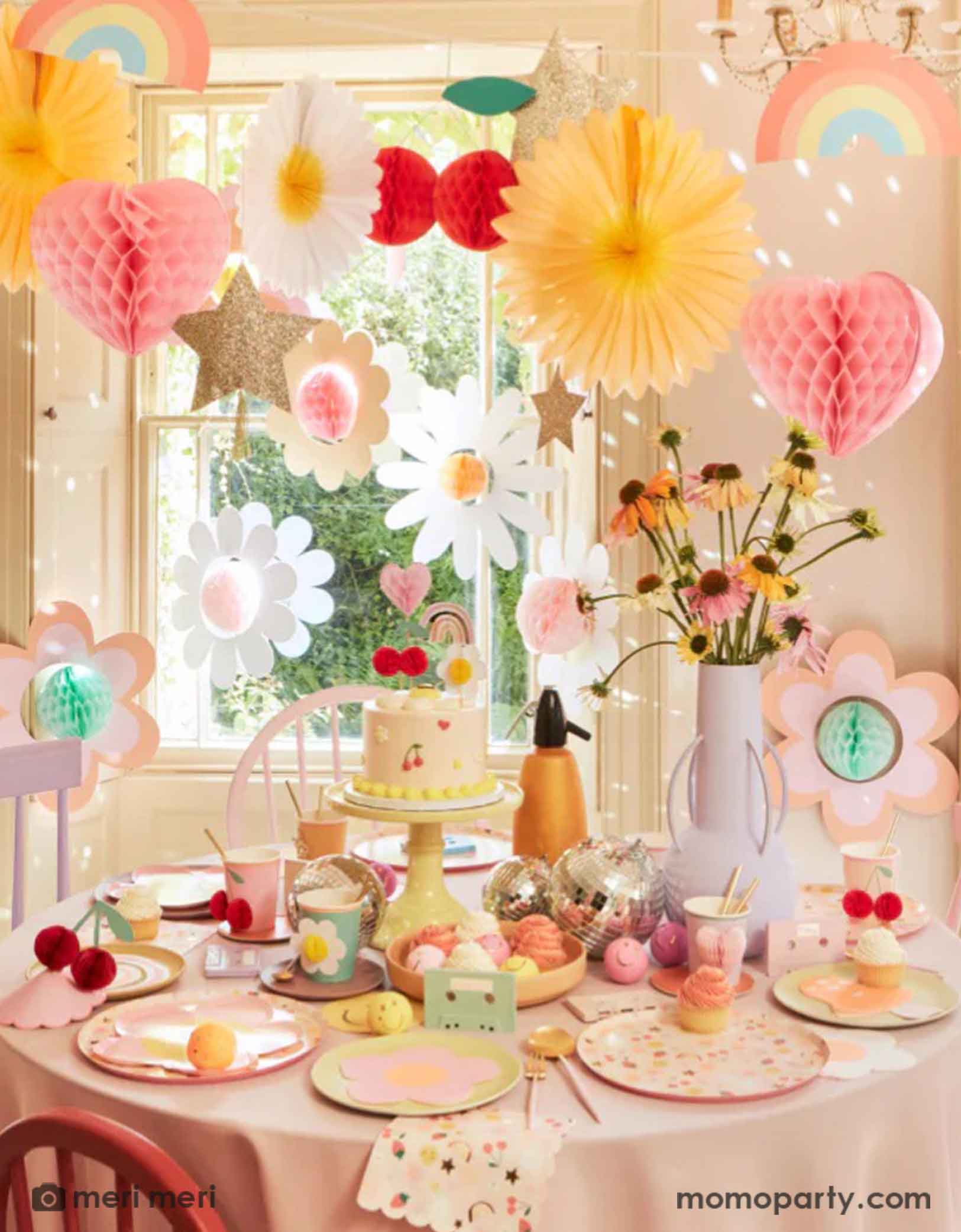 A 90's inspired party table featuring Momo Party's Happy Face Icons party collection by Meri Meri, including happy smiley flower shaped plates, happy icon dinner plates, large napkins and party bags and die-cut napkins in rainbow, smiley face and mushroom shape. Above the table there's a garland with 3D honeycomb decorations of hearts, daisies, rainbows, giving this table a nostalgic and groovy vibe. Perfect for any happy occasion!
