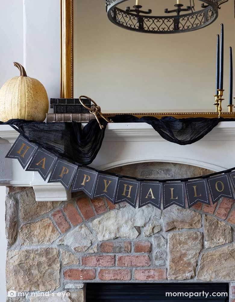 A close up shot of Momo Party's Happy Halloween banner hung over a Halloween mantel that's decorated with elegant Halloween decorations including a black and gold chandelier, black and gold candle holders, a beige colored pumpkin and a gold framed vintage mirror.