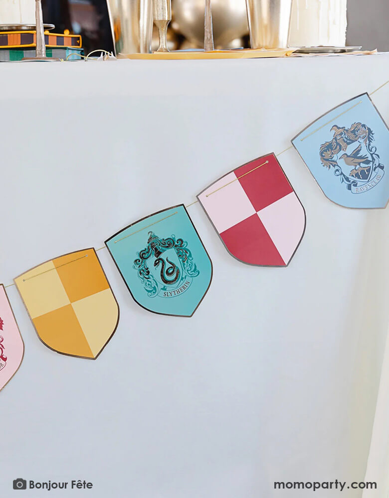 A close up shot of A Harry Potter themed party table adorned with Momo Party's Harry Potter school pride multicolor party banner featuring different Harry Potter house symbols and colors including Hufflepuff, Slytherin, Gryffindor and Ravenclaw. On the party table there are Harry Potter themed party tableware including plates, napkins, party hats, gold cups and white and gold candle sticks.
