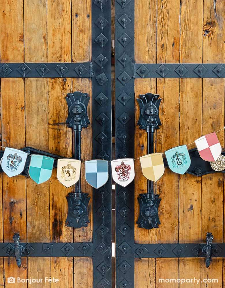 A big Harry Potter Hogwarts-like castle gate adorned with Momo Party's Harry Potter school pride multicolor party banner by Bonjour Fete featuring different Harry Potter house symbols and colors including Hufflepuff, Slytherin, Gryffindor and Ravenclaw - a perfect way to set the scene for a magical Harry Potter themed celebration, be it a kid's birthday party or a Halloween gathering.