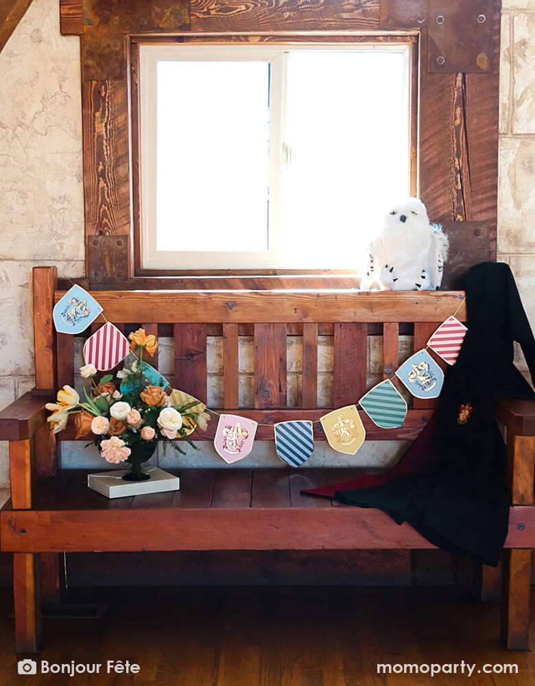 An old, medieval-like wooden bench adorned with Momo Party's Harry Potter school pride multicolor party banner featuring different Harry Potter house symbols and colors including Hufflepuff, Slytherin, Gryffindor and Ravenclaw. On the bench there's a white owl stuffy and a black Harry Potter robe hung across. With a beautiful flower arrangement, it makes a perfect idea for a welcome station that sets the scene for a Harry Potter themed celebration, be it a kid's birthday party or a Halloween gathering.