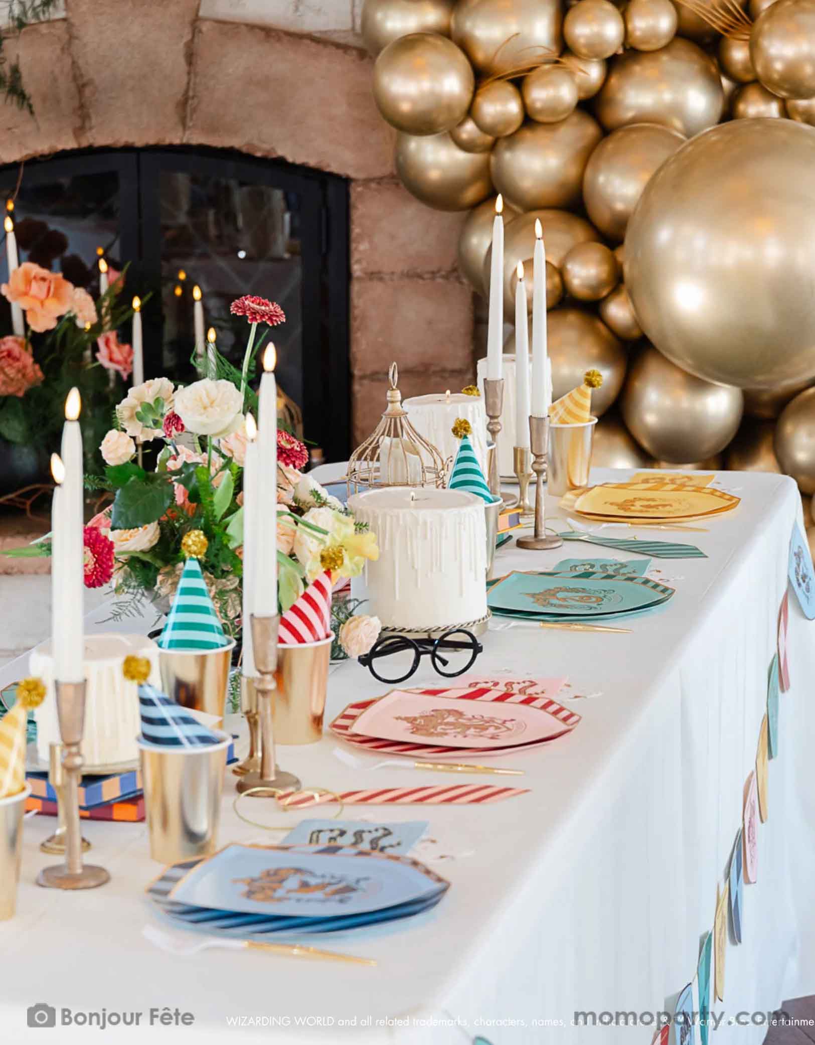 A beautiful Harry Potter themed party table setting featuring Momo Party's Harry Potter house pride shield-shaped 7" side plates with house logos and colors paired with 10.25 striped dinner plates and birthday party hats. In the center of the table there are some candle shaped birthday cakes, bird cages for owls, and some floral arrangement, plus a pair of Harry Potter's glasses. In the back there's a fire placed decorated with chrome gold balloon garland, making this a magical party for kids!
