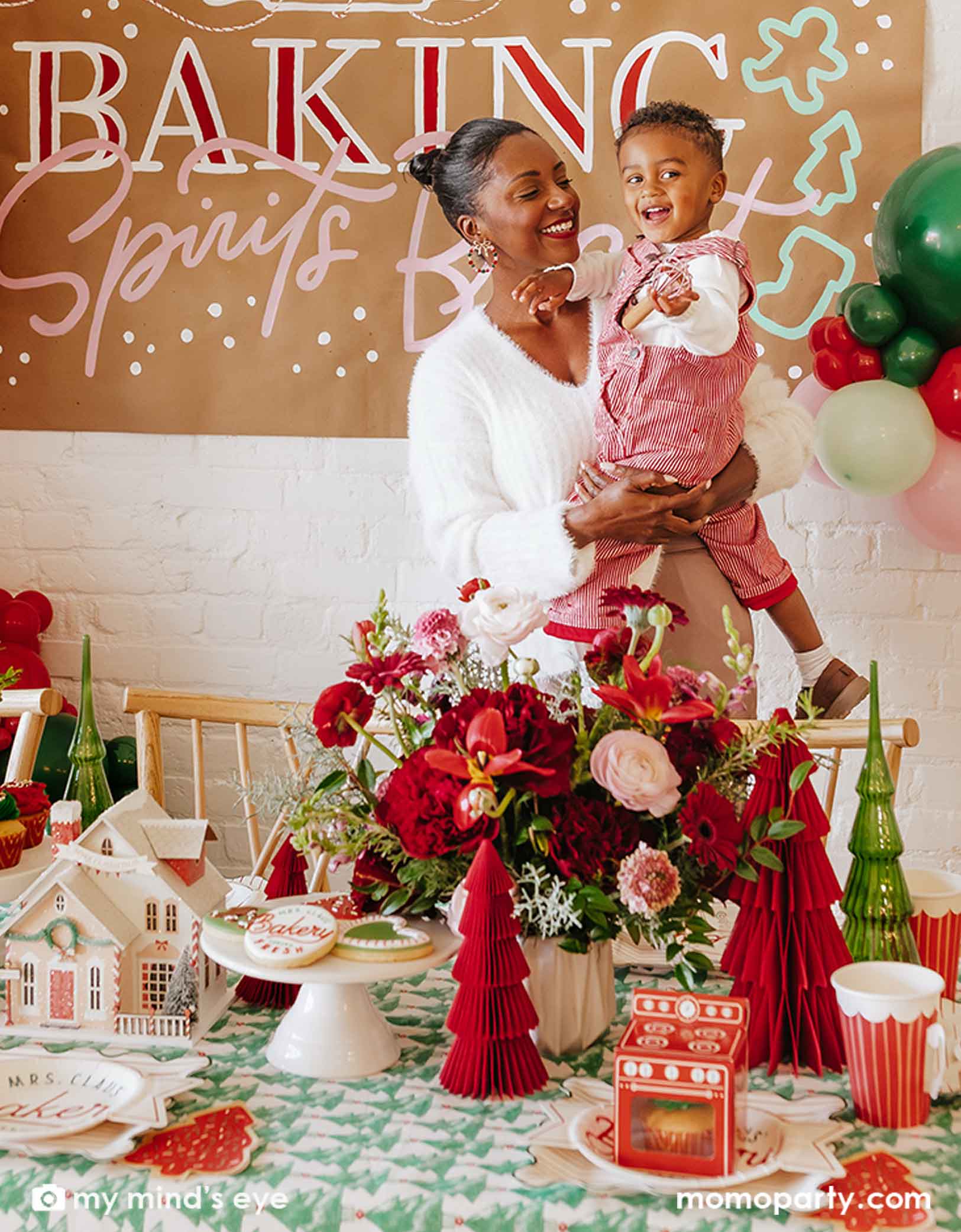 A mom holding a toddler-aged boy in festive Holiday themed outfits standing front of a 'Baking Spirits' sign in a Christmas baking party set up with a table filed with Holiday baking party supplies and decorations from Momo Party including a festive Holiday balloon garland, paper Christmas tree honeycomb decorations, party plates, cups, napkins and more.