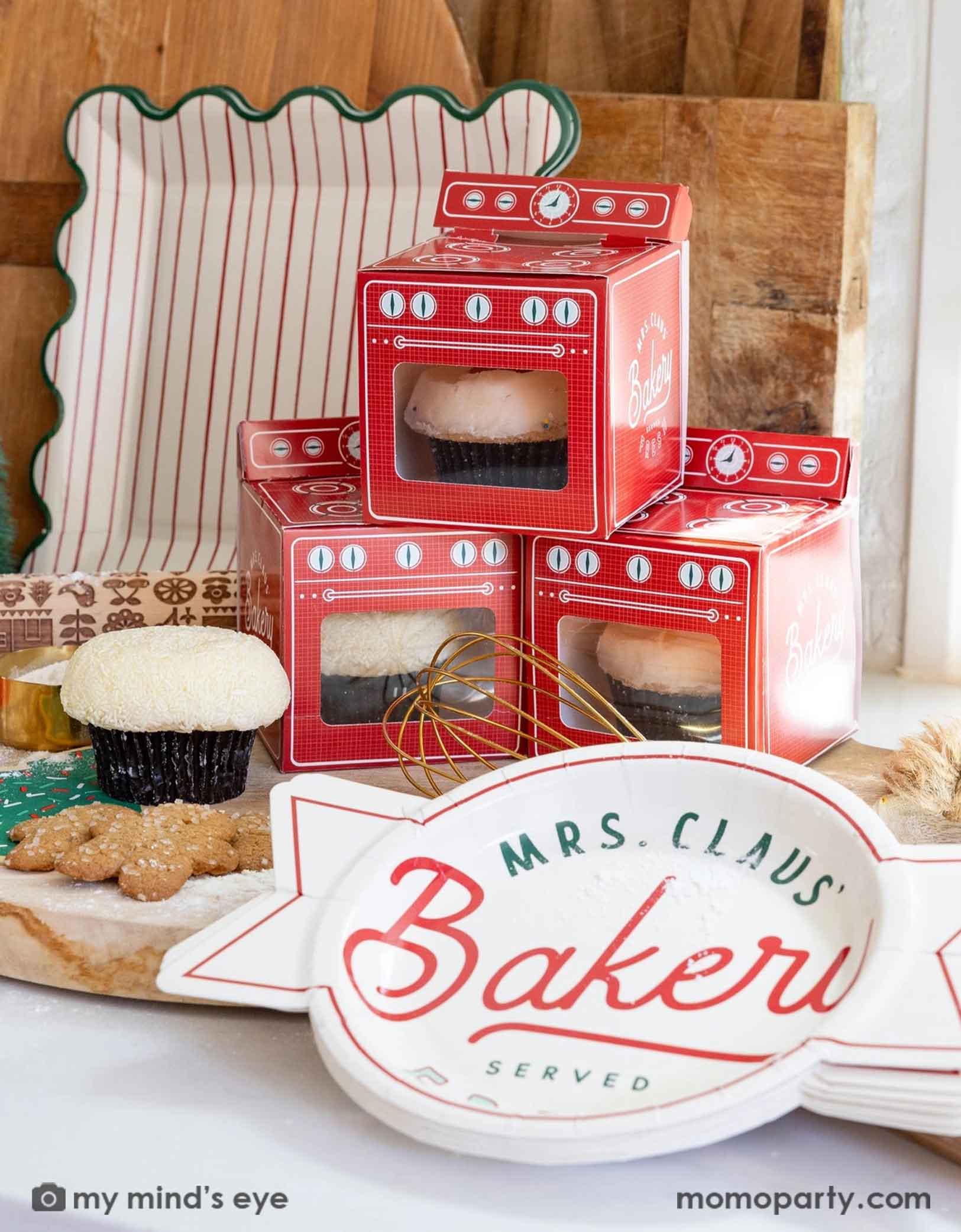 A kitchen counter with Momo Party's Mrs. Claus Bakery party collection featuring the red style oven shaped treat boxes with cupcakes in them. The bakery sign shaped plates, and the red striped plates with green scallop edge. Around the tablewares are some baking utensils including a cutting board, a roller pin, a whisk with some flour around - a cute Holiday baking collection perfectly for kids!