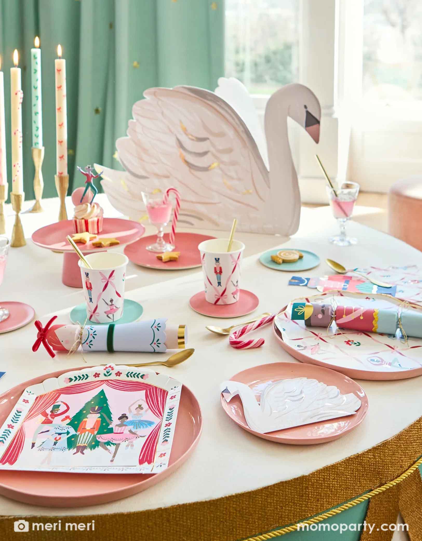 A Holiday children's party table setting featuring multiple Nutcracker themed party supplies from Momo Party including with the Nutcracker stage plates, dinner plates, party cups, napkins, Nutcracker soldier shaped napkin, cupcakes and party crackers, along with swan shaped napkins and centerpiece and pastel long candles on the table, this makes a festive yet elegant table setting for the Holiday season.