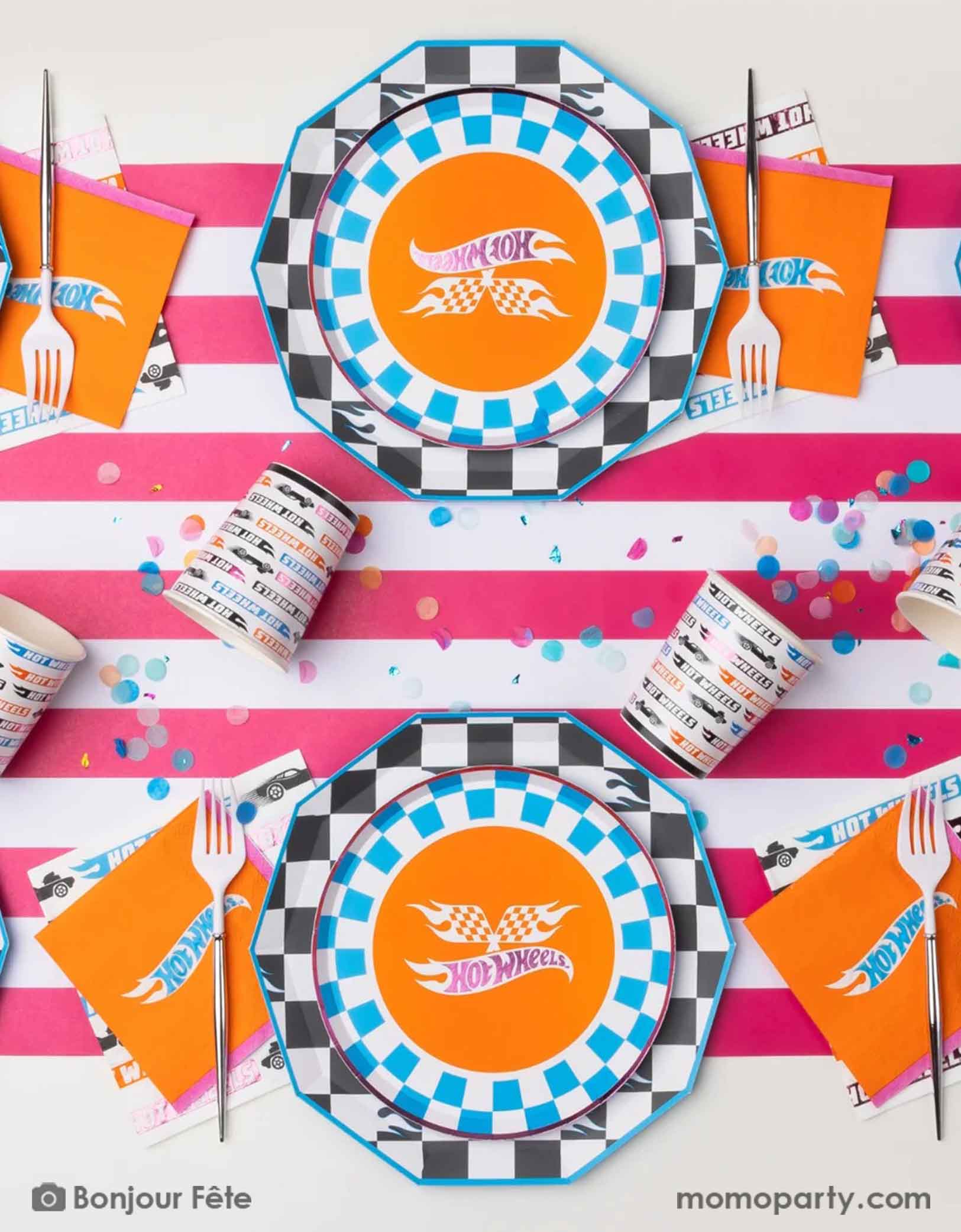 An overhead shot of a race car Hot Wheels inspired party table setting featuring Hot Wheels themed tableware from Momo Party including the checkered dinner plates, the blue checkered Hot Wheels side plates, the orange Hot Wheels small napkins, the Hot Wheels colorful large napkins and matching party cups on a striped pink and white table runner with colorful confetti. A fun and festive party table for kid's race car themed birthday party.