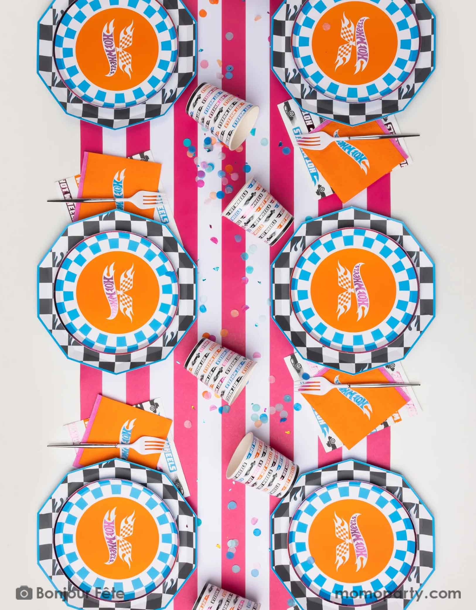 An overhead shot of a race car Hot Wheels inspired party table setting featuring Hot Wheels themed tableware from Momo Party including the checkered dinner plates, the blue checkered Hot Wheels side plates, the orange Hot Wheels small napkins, the Hot Wheels colorful large napkins and matching party cups on a striped pink and white table runner with colorful confetti. A fun and festive party table for kid's race car themed birthday party.