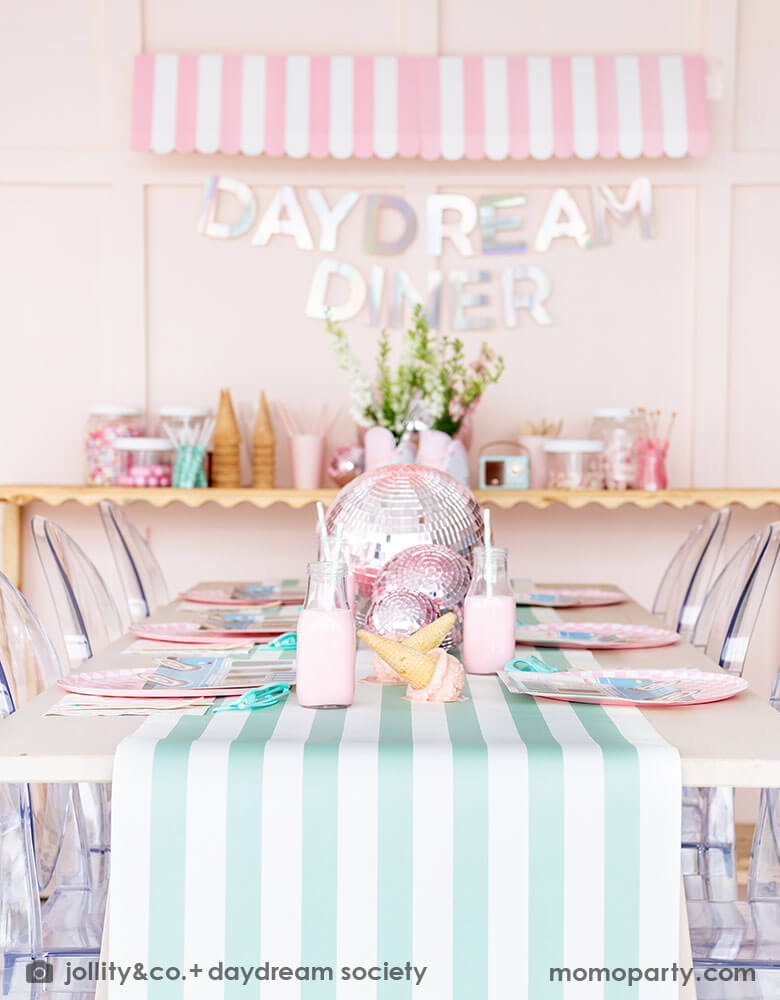 An adorable kid's ice cream themed birthday party tablescape featuring Momo Party's ice cream truck shaped plate, the pink checkered plate, the ice cream icon napkin and pink wooden cutlery on a mint striped table runner. Behind the table there's a sundae station with sprinkle toppings, ice cream cones, adorned with a retro pink striped awning as a small ice cream shop, making this a cute, fun and festive ice cream party set up for kids birthday celebration.