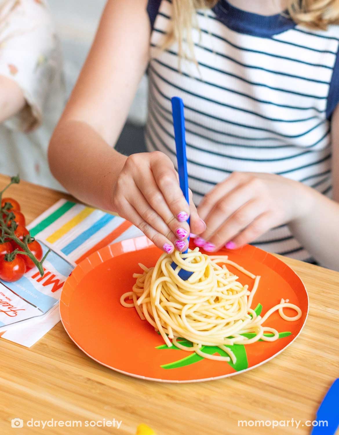 Child's hand with a blue plastic fork in spaghetti on Momo Party's tomato shaped plate with green leaves.