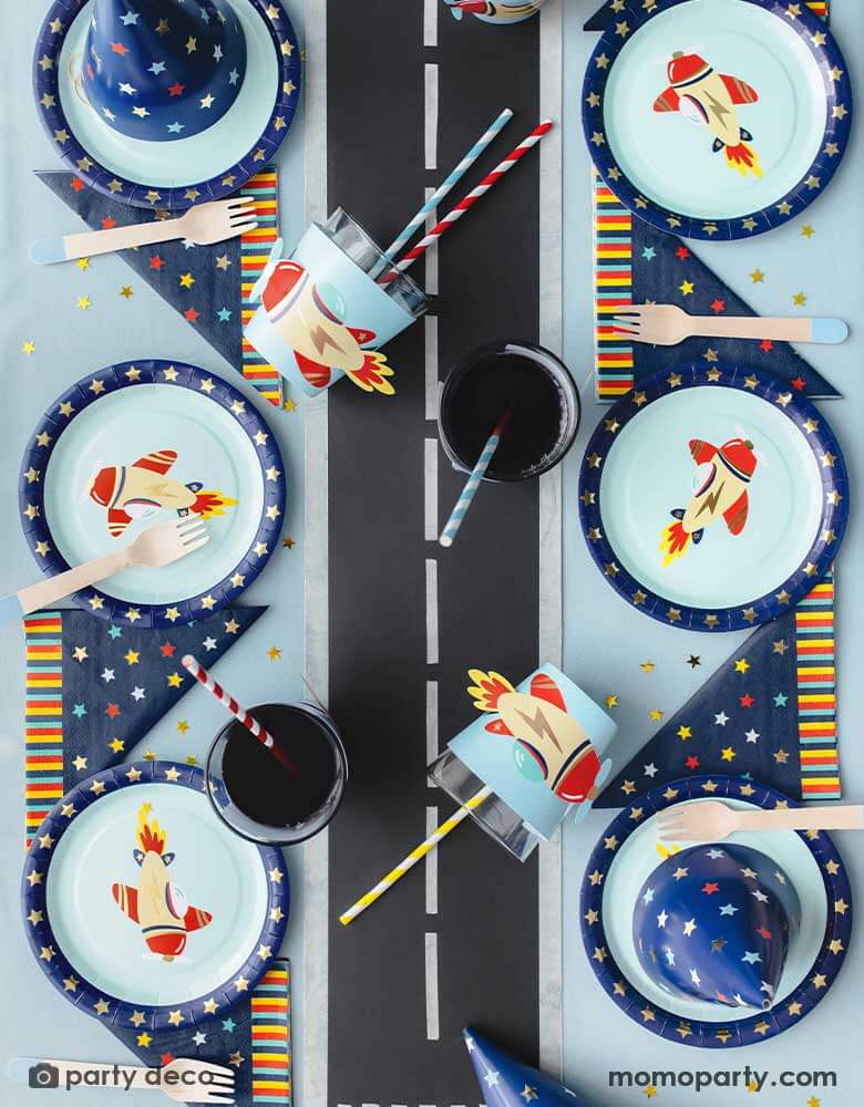 A top view shot of kid's airplane themed party table featuring Momo Party's 7" vintage airplane round paper plates, navy and star around large napkins, vintage airplane cup sleeves wrapped around party cups with red and blue striped paper straws. In the middle of the table is airplane runway track,  making this an adorable inspo for kid's aviation themed birthday party table setting.
