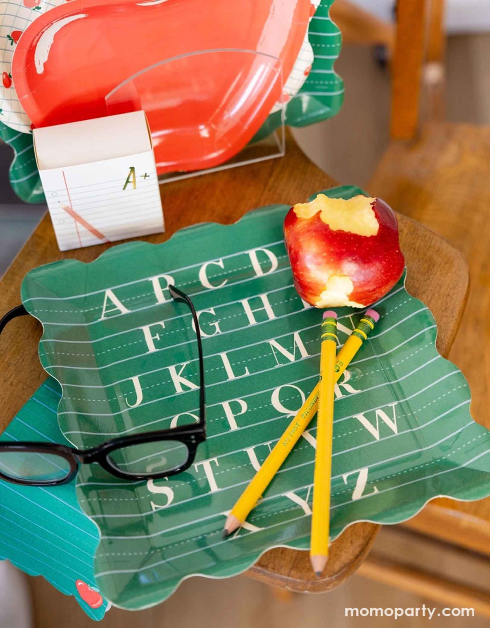 A back to school themed party table placeset featuring various school themed party supplies from Momo Party including the alphabet green large plate with matching apple dinner napkin, along with apple shaped side plate, apple patterned scalloped plate and a notebook shaped treat box. On the alphabet plate there's a half-eaten apple and pair of glasses all on a wooden school chair.