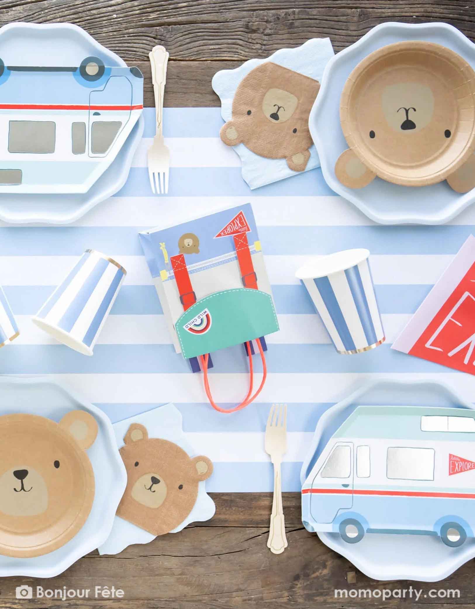 An adorable boy camping party table set featuring multiple camping themed tableware from Momo Party including the bear shaped plates and napkins, camping van shaped plates and a backpack shaped favor bag by Meri Meri from Momo Party. With blue striped party cups and matching paper table runner on top of a wooden picnic table, this gives a perfect inspiration idea for a cute kid's camping themed birthday celebration, be it a "One Happy Camper" or a "Wild One" first birthday party.