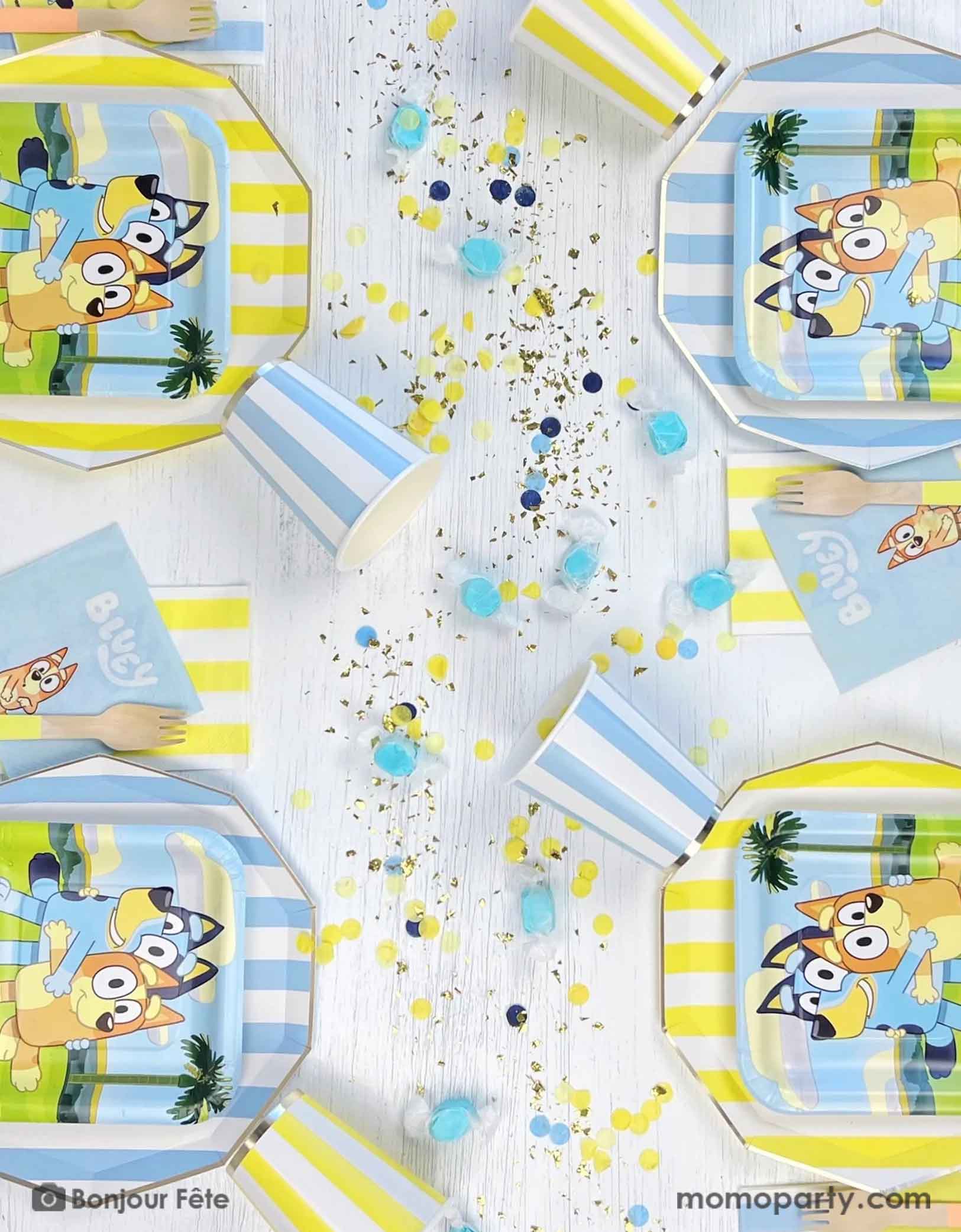 An overhead shot of a Bluey themed kid's birthday party table featuring Momo Party's modern blue and yellow cabana striped tablewares including paper plates, paper cups and napkins. On the dinner plates there are square side plates and party napkins featuring Bluey charcters including Bluey and Bingo, with matching colored confetti and blue colored candies on the table.