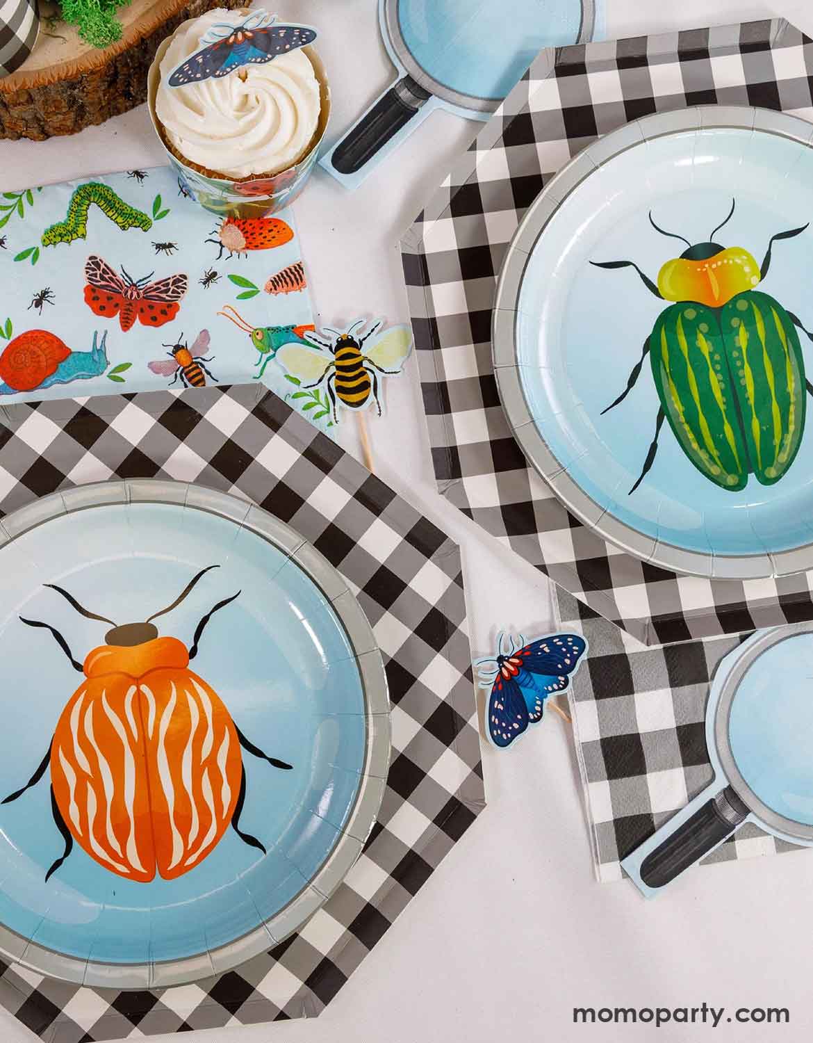 An overview shot of children's bug themed party table setting with bug themed party supplies from Momo Party including two beetle round paper plates on checkered hexagonal dinner plates, napkins with insect pattern, magnifying glass shaped napkins, and insect themed cupcake toppers on a white table.