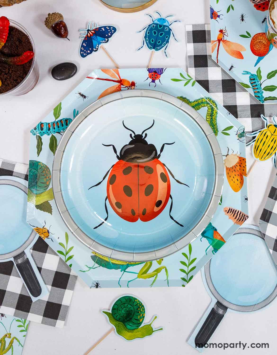 An overview shot of children's bug themed party table setting with a ladybug round paper plate on a hexagonal dinner plate with insect pattern, checkered napkins, magnifying glass shaped napkins, and insect themed cupcake toppers on a white table.