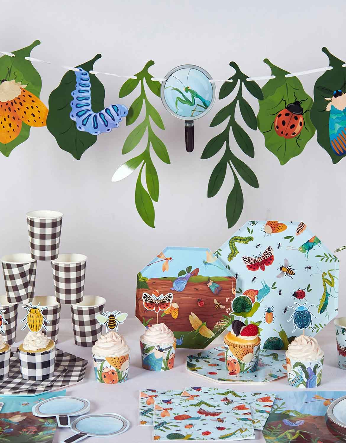 A children's bugs themed party table setting with insect-themed tablewares and decorations with natural design elements from Momo Party, including paper plates, cups, napkins, party banner, cupcake toppers and wrappers.