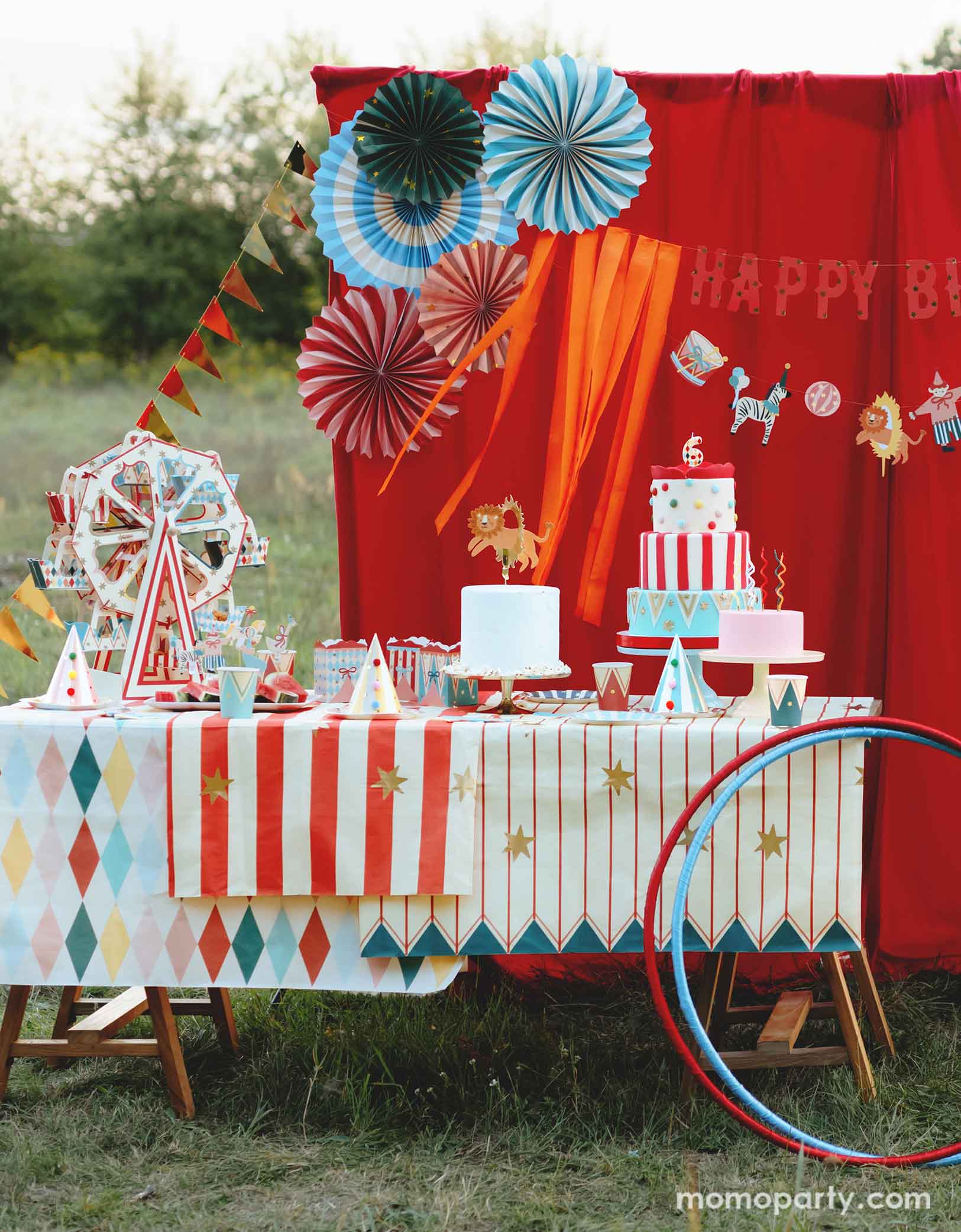 An adorable circus & carnival themed birthday party set up features various circus themed party supplies from Momo Party, including a stunning ferris wheel cupcake stand centerpiece with circus-inspired design, party plates, hats, cups, treat boxes and multiple birthday cakes on a carnival themed tablecloth. In the back there's a red curtain as a party backdrop decorated with circus birthday banner with circus animal party pennants and colorful party fans. A perfect inspo for a fun circus party for kids.