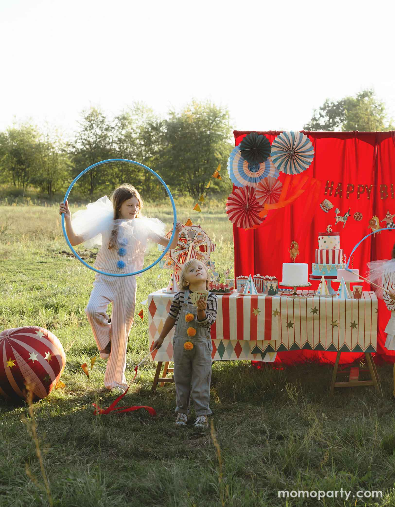 A vintage circus themed party set up in the park featuring various carnival circus themed party decorations from Momo Party including the red striped globe foil balloon next to the red curtain party backdrop. In the front there's a long table with goodies covered with circus themed table covers. A couple of kids dressed in vintage carnival themed clothing playing hula loops, making this a whimsical and playful setting for a fun kid's circus party.
