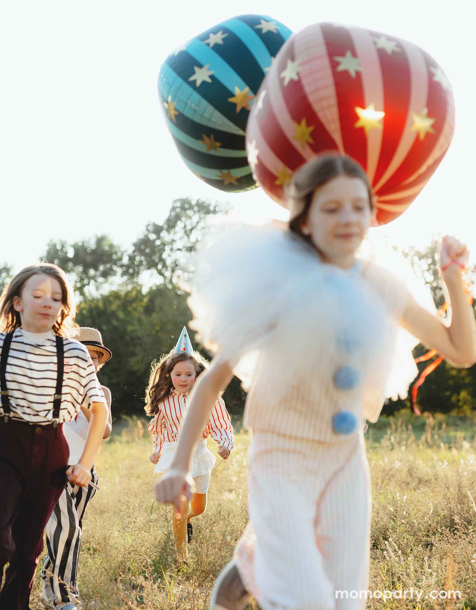 A bunch of kids dressed in quirky vintage circus inspired clothing holding Momo Party's circus themed party balloons running towards the camera. The foil balloons floating in the air include a red globe foil balloon, a green striped globe foil balloon and some gold star foil balloons, perfect decorations for a fun kid's circus/carnival themed celebration.