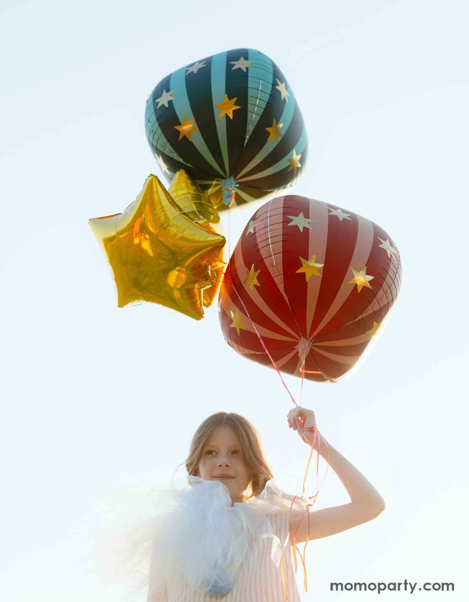A girl dressed in quirky vintage circus inspired clothing holding Momo Party's circus themed party balloons. The foil balloons floating in the air include a red globe foil balloon, a green striped globe foil balloon and some gold star foil balloons, perfect decorations for a fun kid's circus/carnival themed celebration.