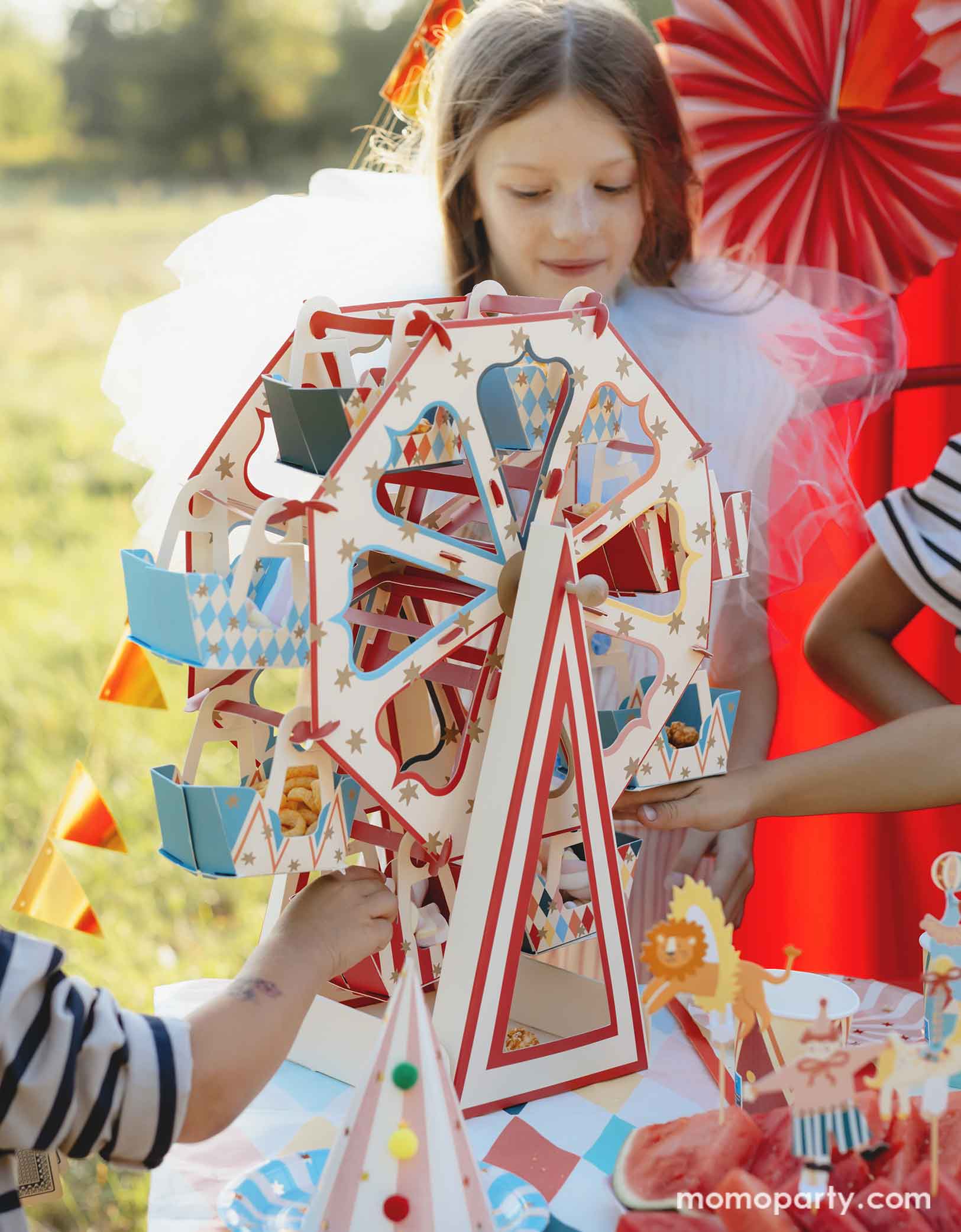 A few kids gathering at the party table in a circus & carnival themed birthday party which features Momo Party's 18.9" x 19.7" Carnival Ferris Wheel Snack Stand by Party Deco. This stunning centerpiece with its circus-inspired design is loaded with treats and cupcakes and they kids are grabbing the goodies from this fun treat stand. In the back, there's a red stage curtain as a party backdrop decorated with circus themed birthday banner with circus animal party pennants and colorful party fans.