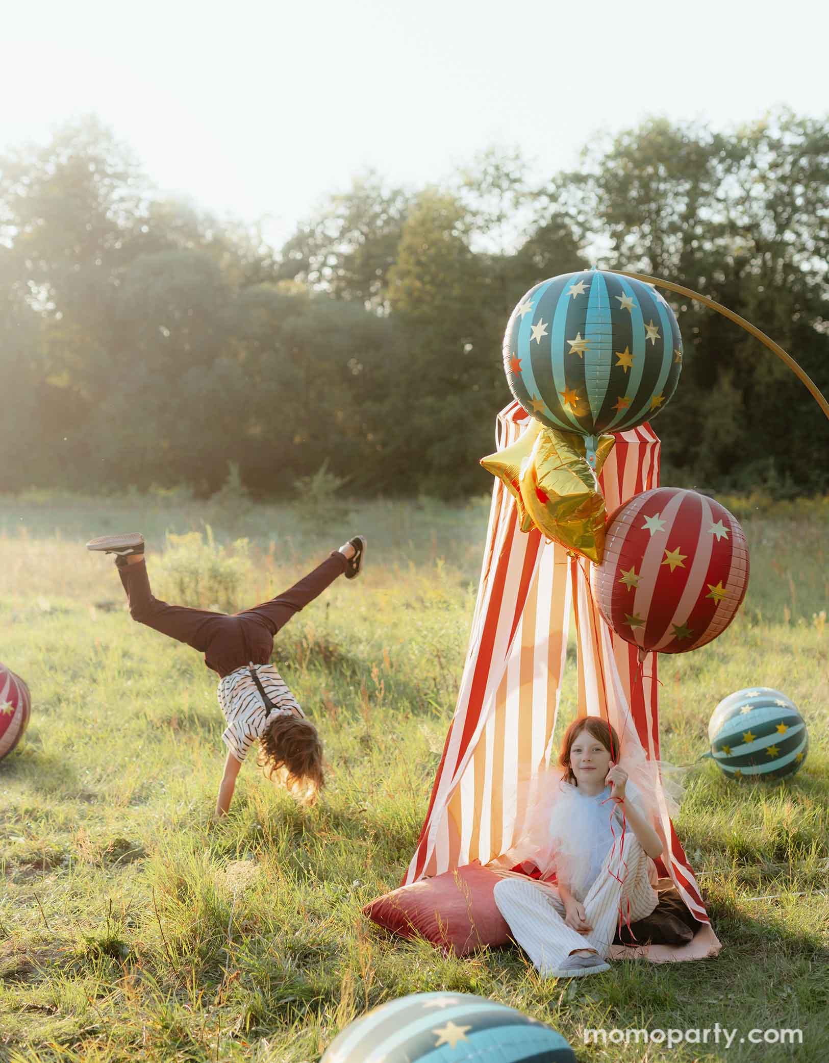 Two kids dressed in vintage and quirky clothing hanging out and playing in a vintage circus themed party featuring various carnival circus themed party decorations from Momo Party including the red striped globe foil balloon, the green striped globe foil balloon and gold star foil balloons floating next to a red and white striped circus themed play tent for kids. With sun dawn lighting from the back, making this a whimsical look for a fun circus/carnival themed celebration.