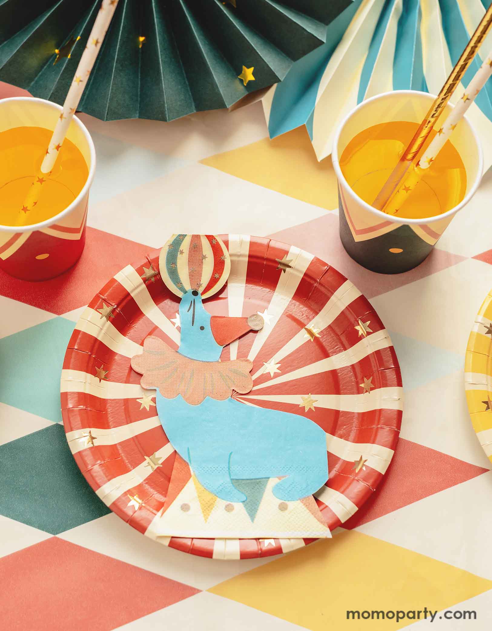 A carnival/circus themed part table featuring Momo Party's circus themed party tableware including a red striped paper plate and a sea lion playing with a ball shaped napkin, with matching party cups with gold foil star paper straws and some festive party fans on a circus themed striped table cover - making this great table setting idea for a fun and festive kid's carnival themed birthday celebration!