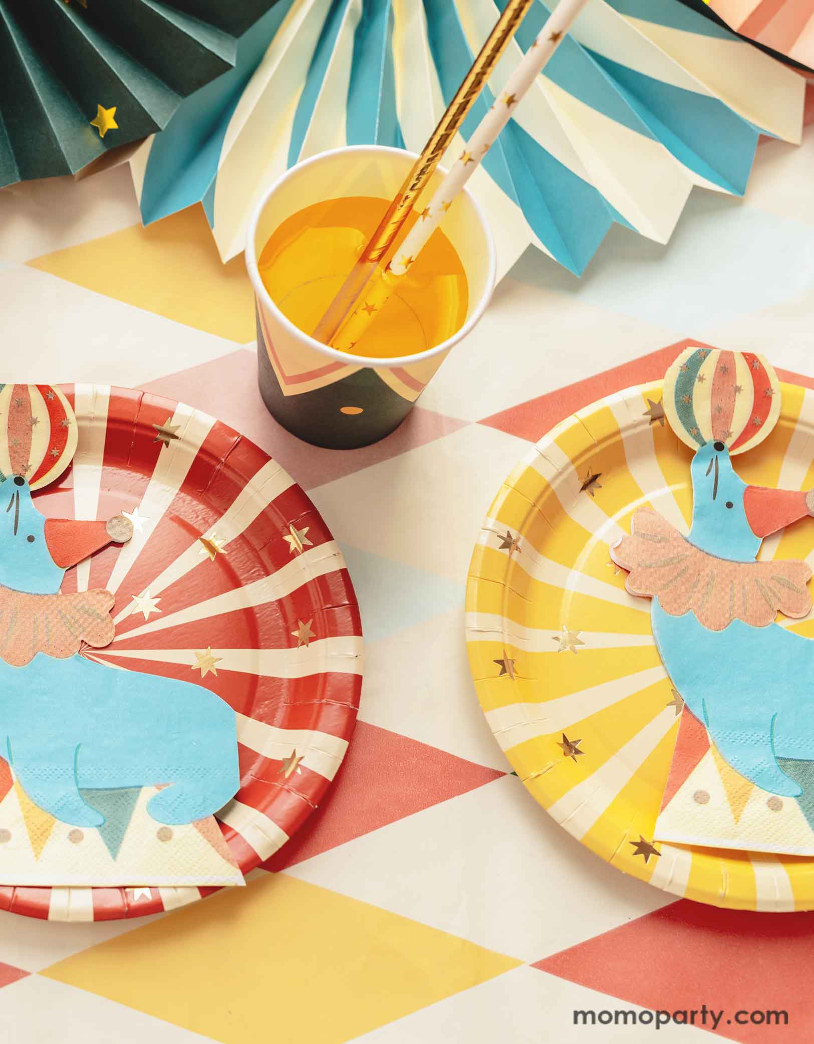A carnival/circus themed part table featuring Momo Party's circus themed party tableware including a red and yellow striped paper plates and sea lion playing with a ball shaped napkins, with matching party cups with gold foil star paper straws and some festive party fans on a circus themed striped table cover - making this great table setting idea for a fun and festive kid's carnival themed birthday celebration!