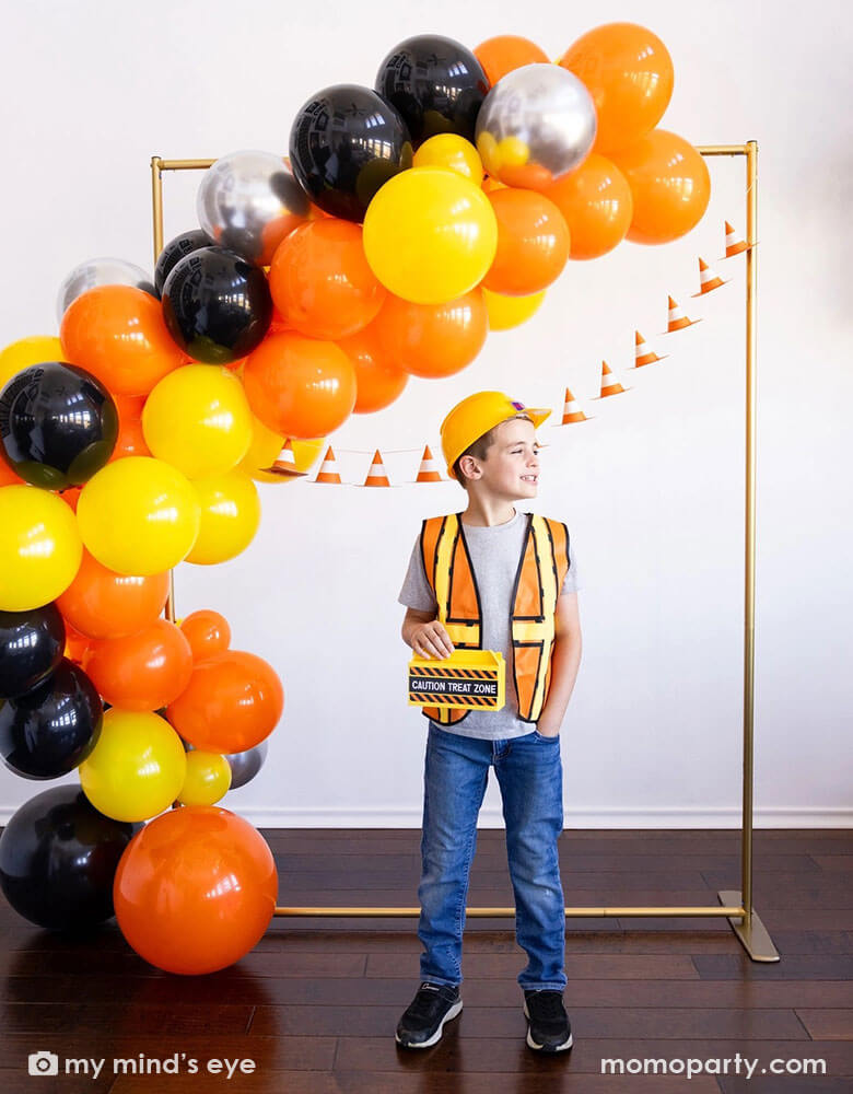 A school aged boy in his construction costume and hard hat holding Momo Party's toolbox shaped treat box by My Mind's Eye standing in front of a festive construction themed party decoration which features a balloon garland in black, orange silver and yellow and a construction cone banner hung over on the wall - a perfect decoration idea for kid's construction builder themed birthday celebration party.
