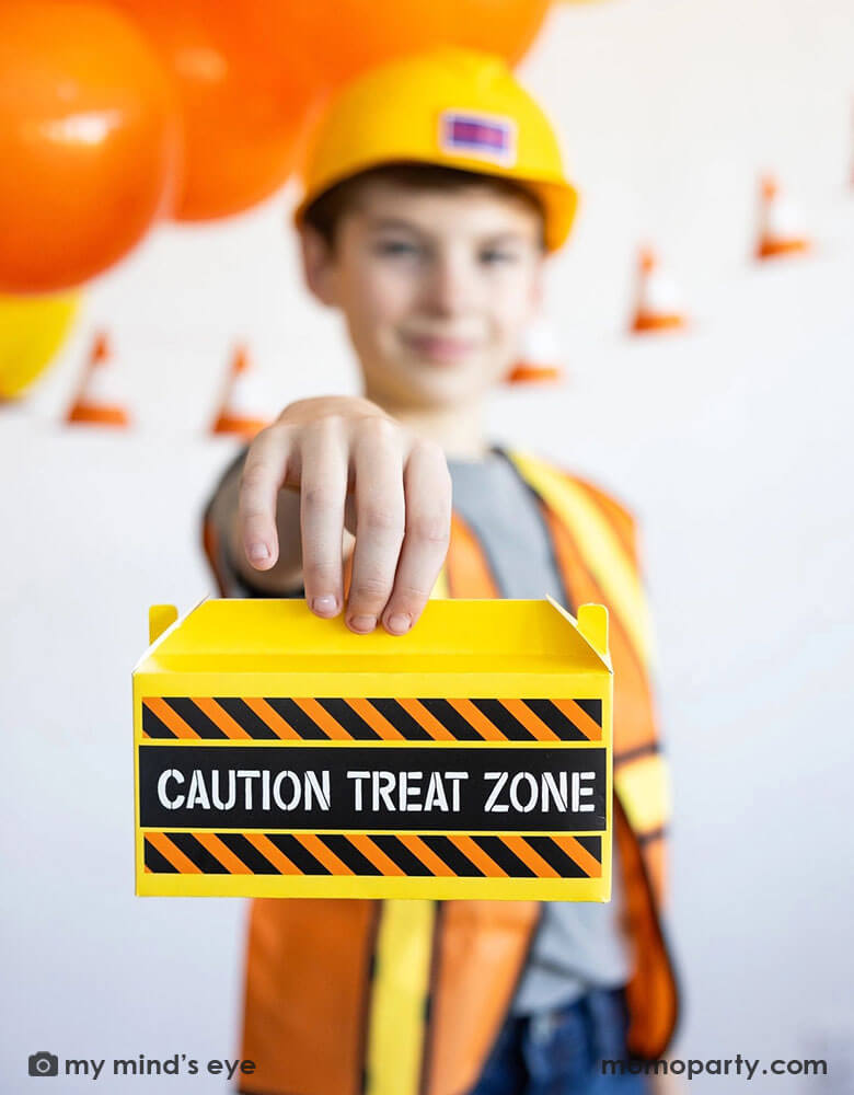 A school aged boy in his construction costume and hard hat holding Momo Party's toolbox shaped treat box by My Mind's Eye standing in front of a festive construction themed party decoration which features a balloon garland in black, orange and yellow and a construction cone banner hung over on the wall - a perfect decoration idea for kid's construction builder themed birthday celebration party.