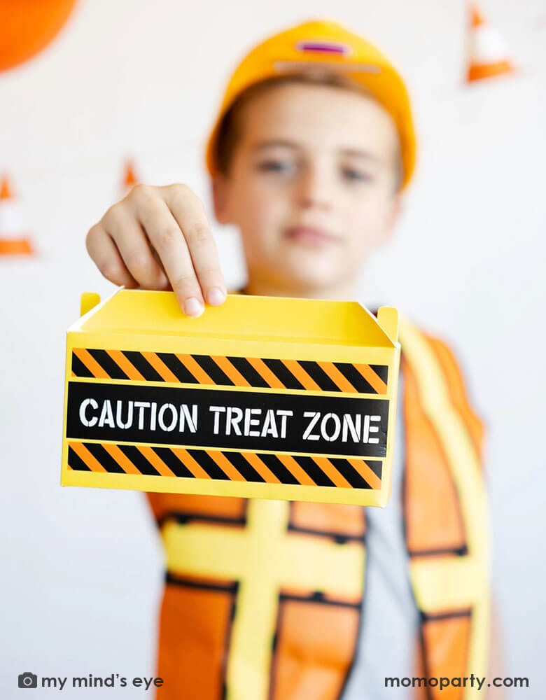A school aged boy in his construction costume and hard hat holding Momo Party's toolbox shaped treat box by My Mind's Eye standing in front of a festive construction themed party decoration which features a balloon garland in black, orange and yellow and a construction cone banner hung over on the wall - a perfect decoration idea for kid's construction builder themed birthday celebration party.
