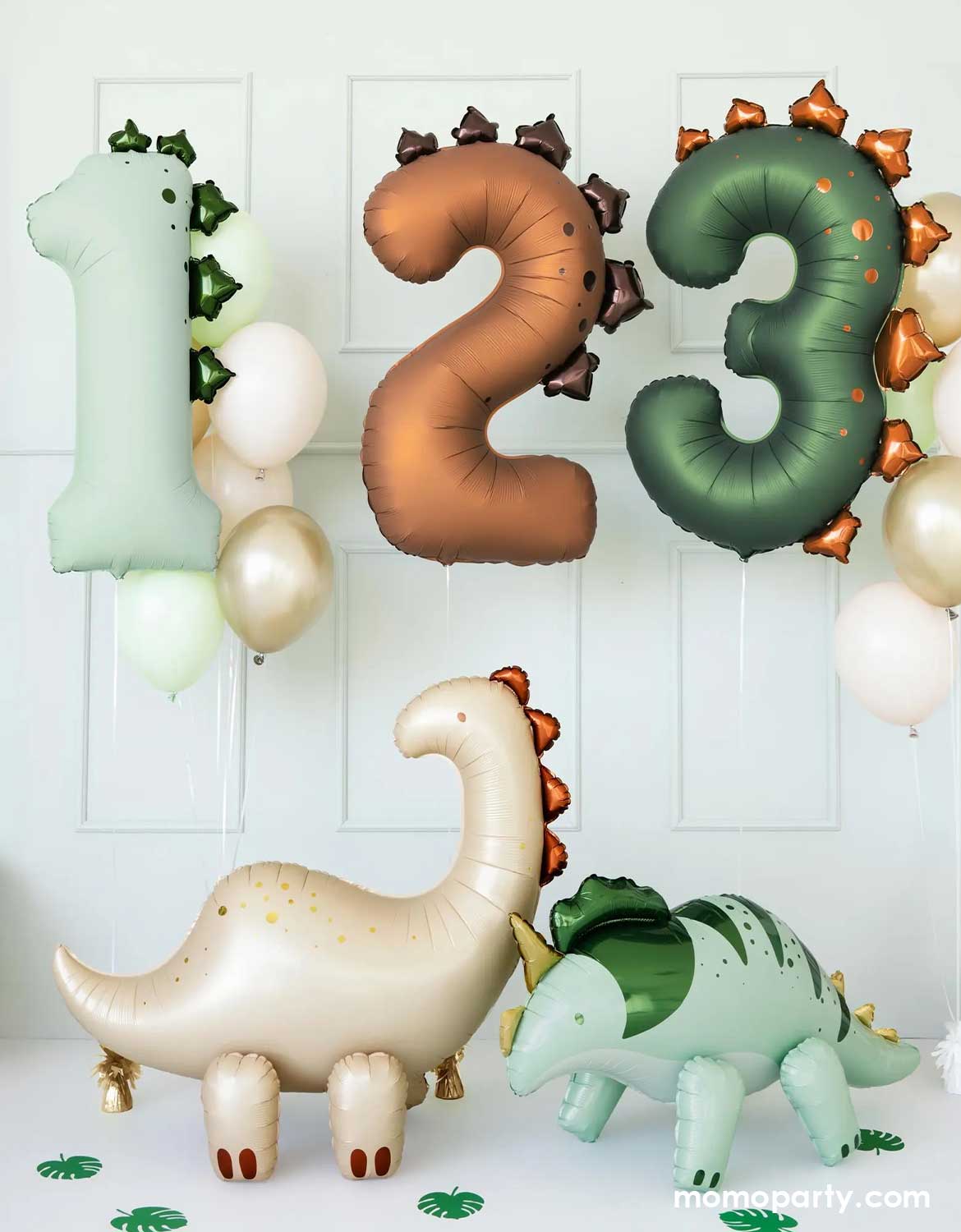 An adorable dinosaur-themed balloon decoration featuring Momo Party's mint number one dinosaur balloon, brown number two balloon and dark green number three balloon along with color-coordinated latex balloons floating against a white wall. On the floor, there are two standing dinosaur shaped foil balloons from Momo Party with palm leaf cutouts around, making this a cute set up for kid's dinosaur themed birthday celebrations.