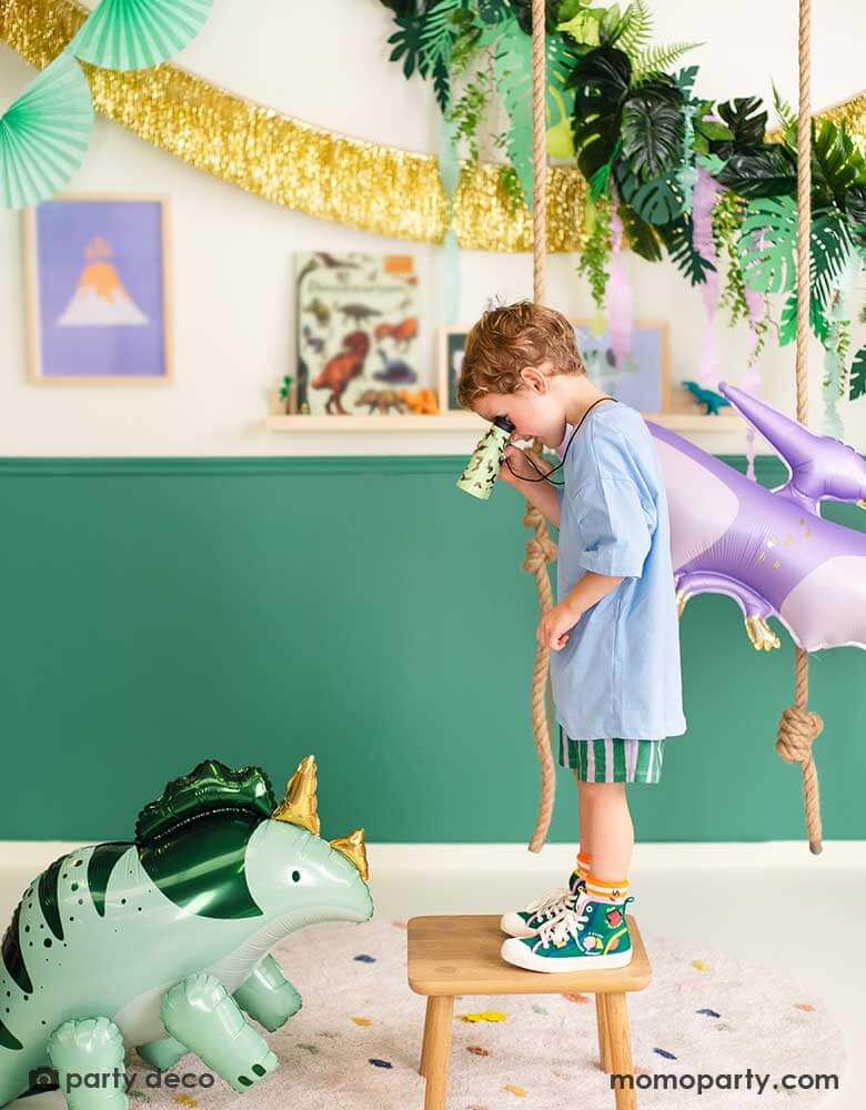 A boy is watching Momo Party's Triceratops shaped foil balloon through his binoculars. He's in a room decorated with prehistorical themed decoration including a lilac PTERODACTYL shaped balloon, a gold tinsel fringe garland, a tropical leaves garland from Momo Party.