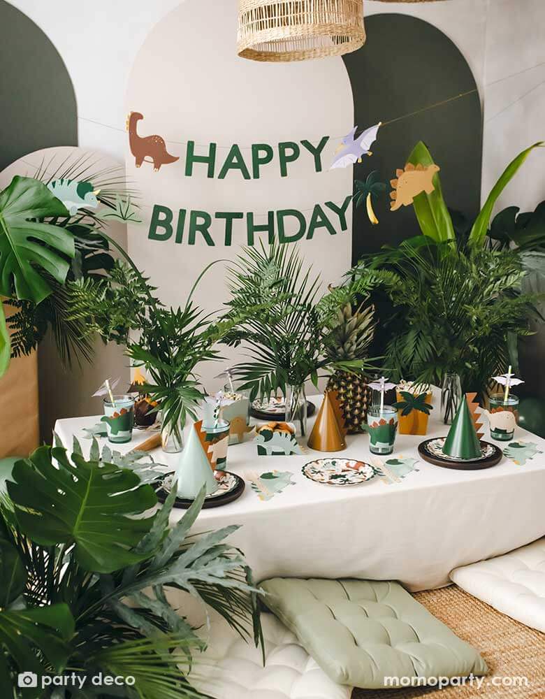 An adorable kid's dinosaur themed party set up featuring Momo Party's dinosaur themed party supplies and decorations by Party Deco including dinosaur paper plates, cup sleeves, dinosaur cake toppers, dinosaur spikes party hats, dinosaur napkins and a dinosaur happy birthday banner hung on the party backdrop wall. With a lot of tropical plants as the centerpiece, it makes a cute and modern inspo for kid's dinosaur themed birthday party.
