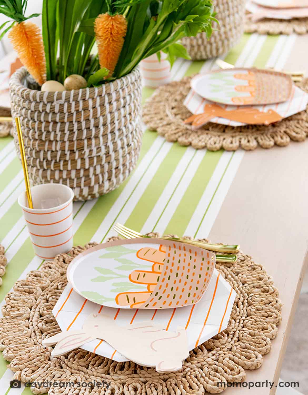 An Easter bunny themed table setting with Momo Party's bunny themed tablewares including carrot basket plates, bunny shaped napkins and matching cups and utensils on a straw-weaved placemats over a green and white striped table runner. In the center there's a green and carrot floral arrangement as the centerpiece.