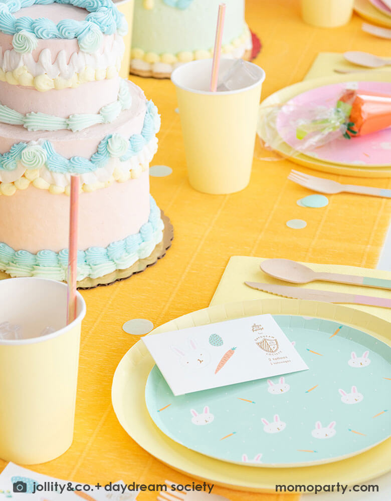 A kid's Easter party table featuring Momo Party's Easter Fun party collection by Daydream Society including Easter Fun plates in mint and pink, Easter Fun temporary tattoos, pale yellow dinner plates and party cups. On the table there are two vintage inspired buttercream cakes with pastel piping, making this a perfect inspiration for Easter brunch hosting occasion!