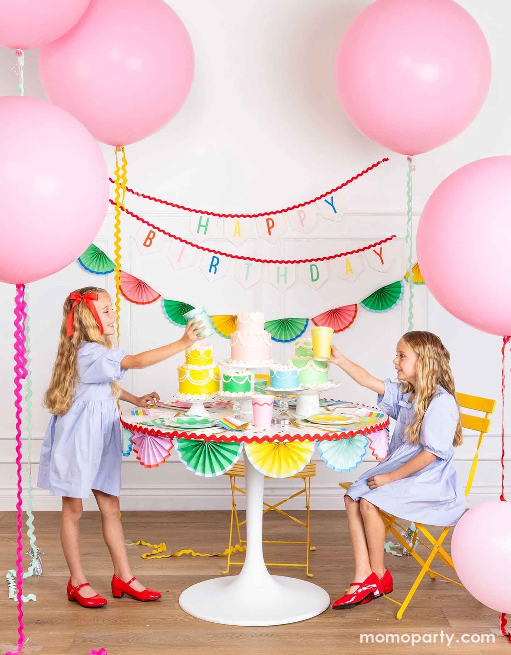 A cheerful and colorful birthday party set up featuring various festive birthday party supplies from Momo Party including rainbow colored tablewares of plates, cups, napkins on the table along with multiple pastel colored birthday cakes. The table is decorated with colorful bunting garland around and in the back of the wall hung a Happy Birthday felt garland and the colorful bunting banner. On the sides are some jumbo pink balloons floating above and two girls sitting at the table and holding cups.