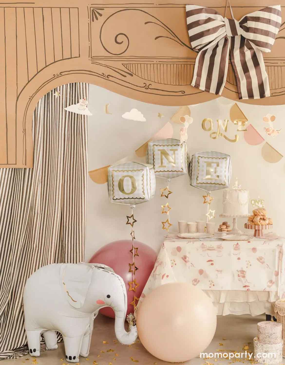 A styled kid's first birthday party setup with a beige arch backdrop, striped curtain, large striped bow, jumbo foil balloons, and Momo Party's standing elephant foil balloon, and a table covered in a pink floral tablecloth with cakes and cupcakes on it.