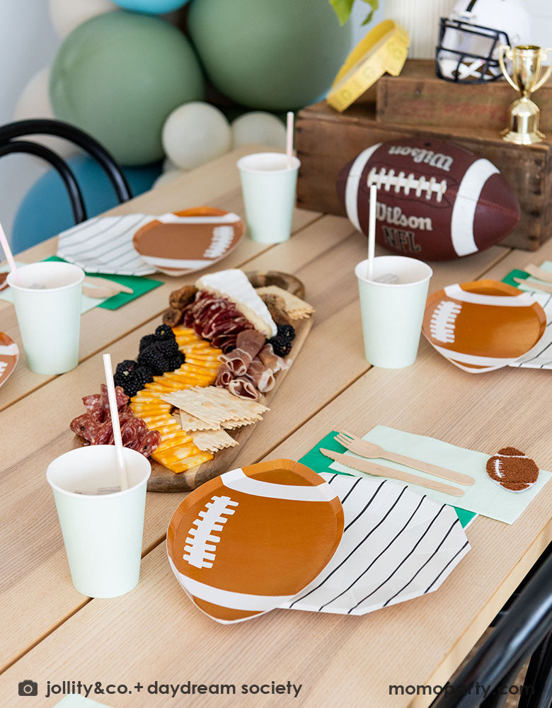 A modern kid's football party table filled with Momo Party's football themed party tableware by Daydream Society including the die-cut football shaped plates, black striped plates, green and mint napkins and party cups. In the center of the table there's a cheese charcuterie board and some decorations like a football, a trophy and a football helmet as the centerpiece - a great inspirations for football themed celebrations!