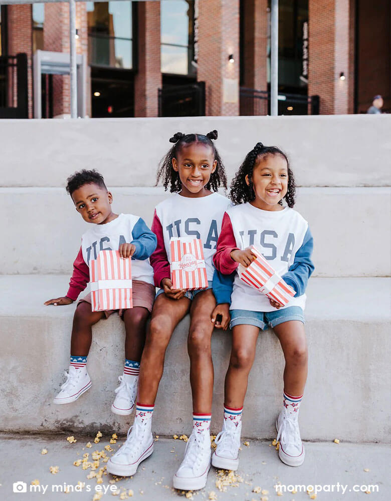 Three children dressed their USA sweatshirt sitting on a bench eating Cracker Jacks of Momo Party's Stars and Stripes Treat Bags by My Mind's Eye featuring red striped design with "Hip Hip Hooray" on it, it's perfect for at this year's Fourth of July parade viewing!