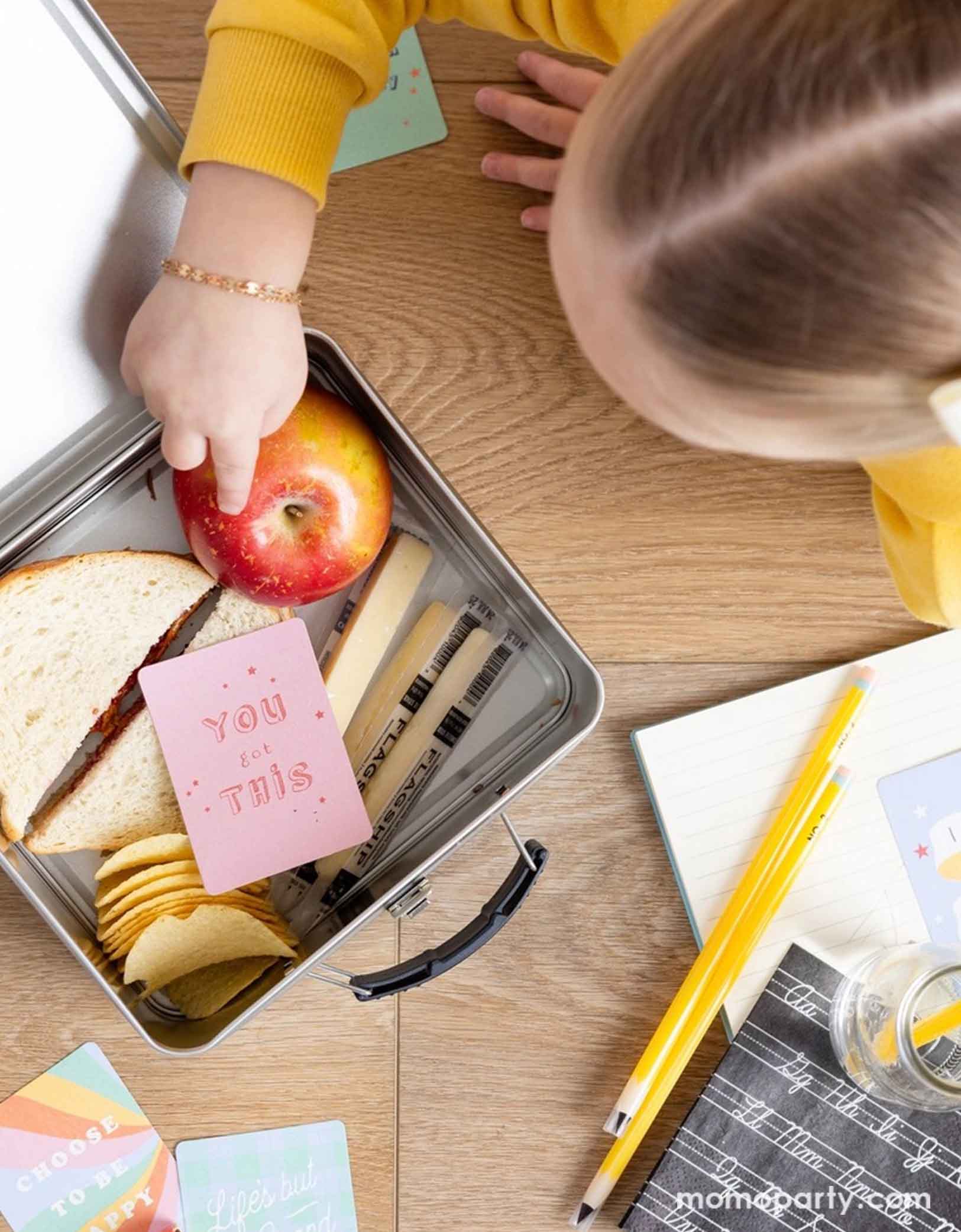 An overhead shot of a girl taking food out from her tin lunch box with a sandwich, some chips, some string cheese and an apple. There's a lunchbox card with "You got this" on it. Next to the lunch box are various school themed party supplies including the reusable pencil straws, cursive alphabet small napkins and some school supplies themed plates.