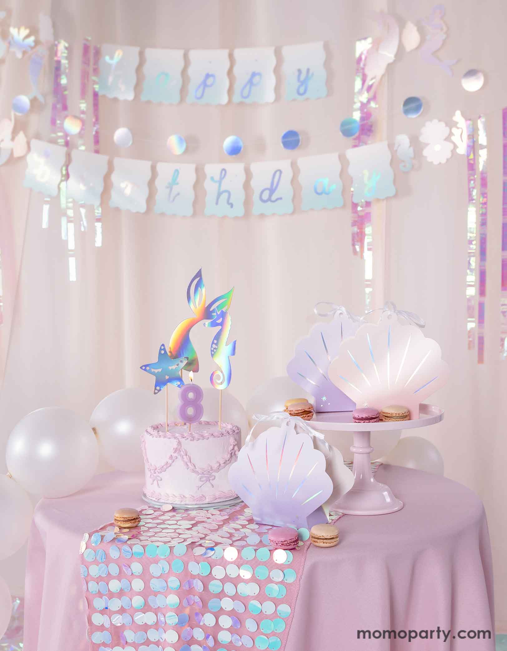 A mermaid birthday party table set uo featuring various mermaid themed party supplies from Momo Party including the ombre pink and lilac shell shaped treat boxes, a pink butter cream cake topped with mermaid themed toppers. Behind the table there's a pink curtain adorned with the mermaid happy birthday party banner as the backdrop. Along with pearl white balloons around, creating a dreamy and whimsical scene for a magical under the sea bash.