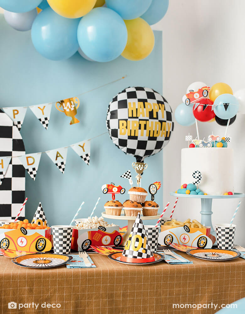 A kid's race car themed party table filled with race car themed party supplies from Momo Party including wheel shaped plates, checkered flag GO! party hats, blue race car large napkin, and checkered party cups. On the table there are muffins topped with race car themed cupcake toppers and vintage race car shaped snack holder filled with popcorn and onion ring chips. Above the table, there's a race car flag themed birthday banner and checkered happy birthday foil balloon.