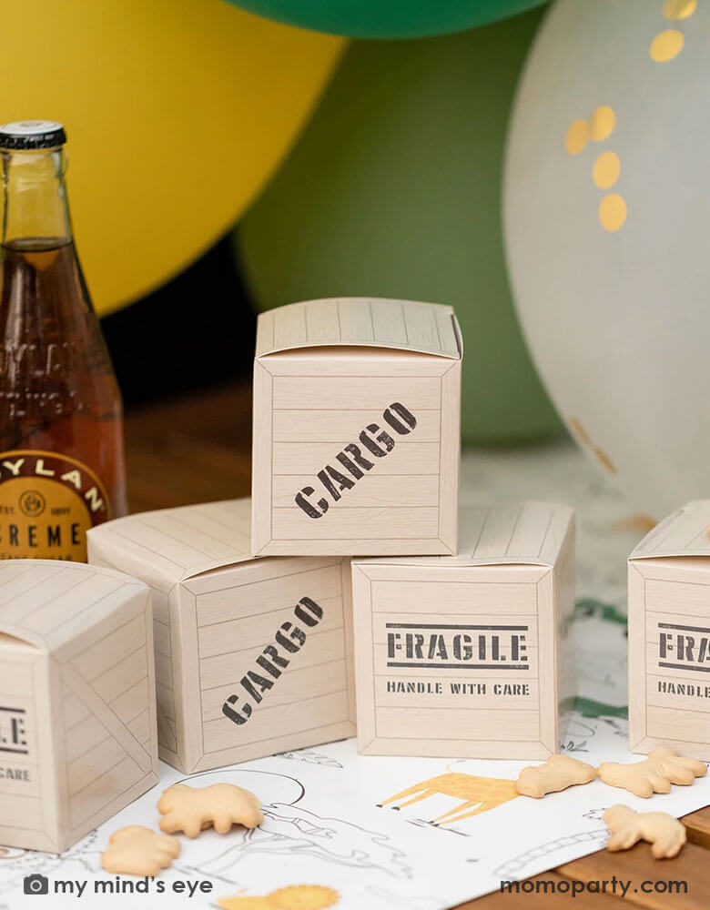 A safari themed party table featuring a few Momo Party presents 3" x 3' safari adventure cargo favor boxes by My Mind's Eye, laid above the safari adventure paper table runner featuring lots of safari animsl. Designed to resemble cargo boxes, each favor box has "Cargo" & "Fragile, handle with care" on the sides. On the party table you can see some soda bottles, animal crackers around the favor boxes and balloons in yellow and green shades, making this a great look for kid's safari themed birthday party.
