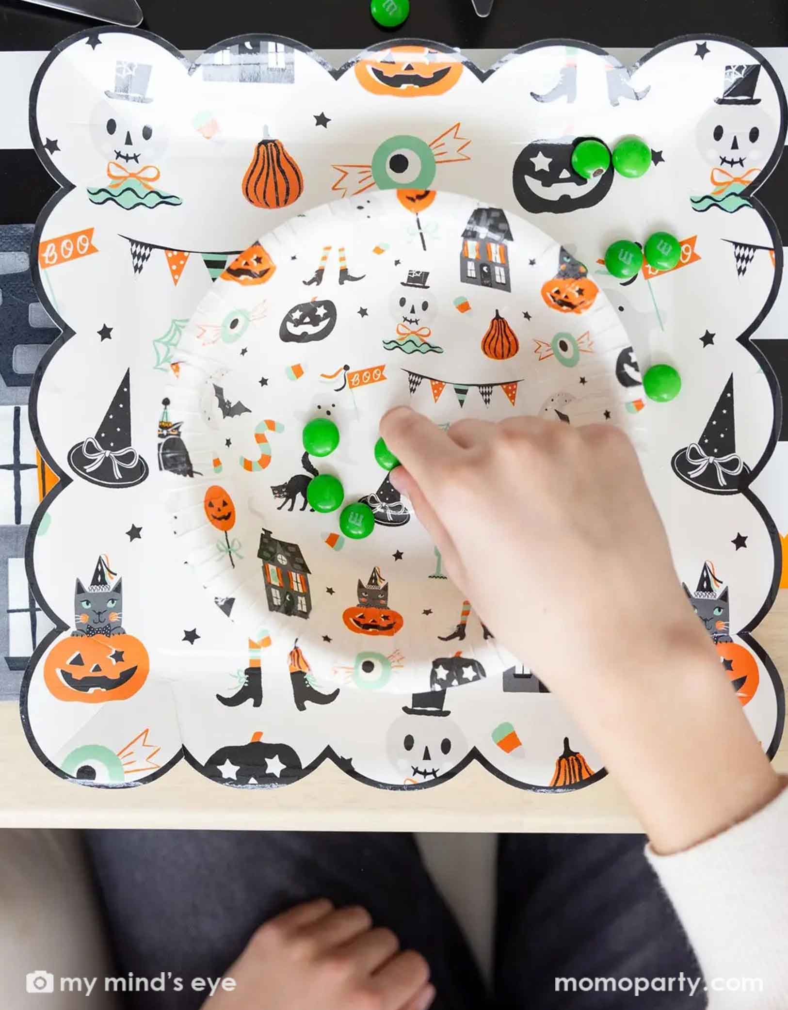 A overhead shot of a festive Halloween-themed table setting with Momo Party's Trick or Treat dinner plates and bowls, the haunted mansion shaped napkins and black cutlery set on a black orange and white striped table runner fro Momo Party. On the plates there are some green candies spread across, featuring a child's hand interacting with them.