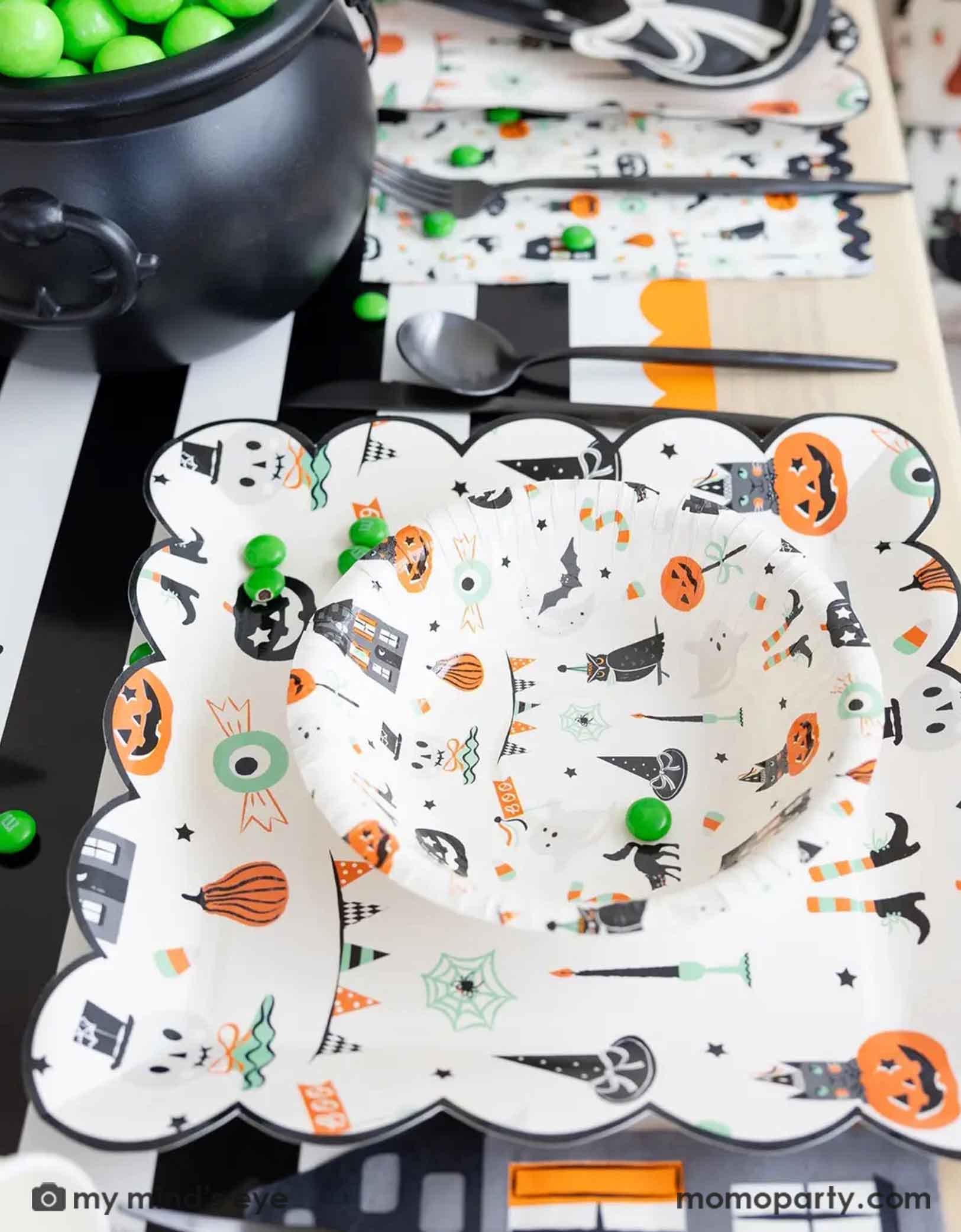 A side shot of a festive Halloween-themed table setting with Momo Party's Trick or Treat dinner plates and bowls, the haunted mansion shaped napkins and black cutlery set on a black orange and white striped table runner fro Momo Party. On the table there's a cauldron filled with green gum balls and some green candies spread across the table. A perfect set up for kid's friendly Halloween celebration.