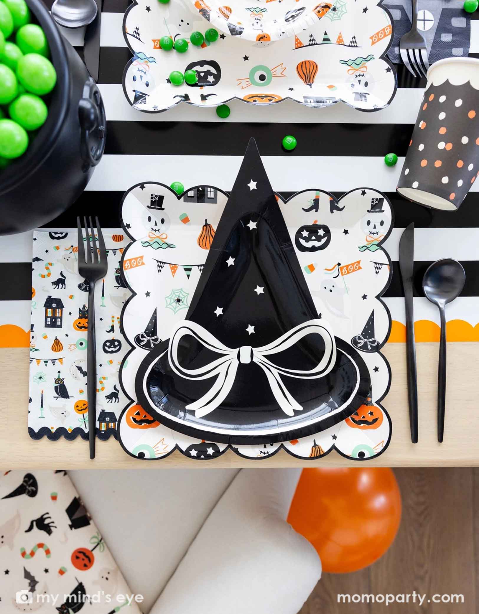 A overhead shot of a festive Halloween-themed table setting with the witch hat shaped plate, Trick or Treat dinner plates and bowls, the haunted mansion shaped napkins and black cutlery set on a black orange and white striped table runner fro Momo Party. On the table there's a cauldron filled with green gum balls and some green candies spread across the table. A perfect set up for kid's friendly Halloween celebration.