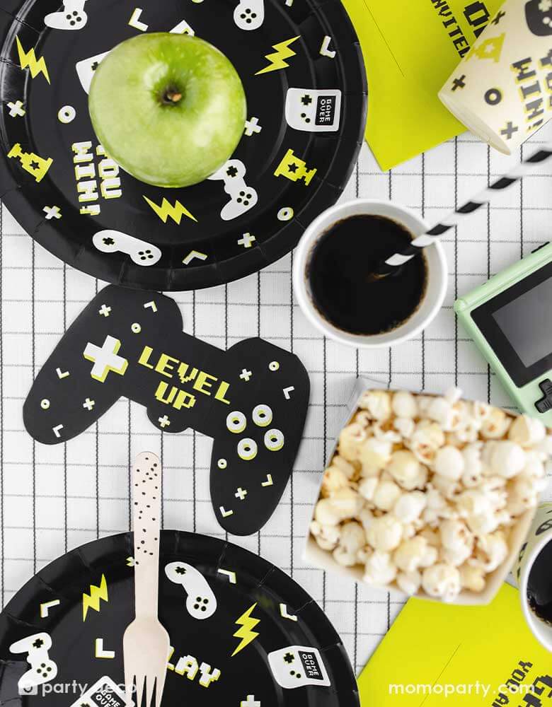 A video game themed party table features Momo Party's black Video Game round party plates with video game devices and controller motifs on them. Next to the plates are a game controller shaped napkins with "LEVEL UP" message on them, with the coordinating video game party cups with black striped paper straws from Momo Party. On the table covered with black and white grid tablecloth, the are some popcorns and an green apple as snacks, ready for a fun game time!