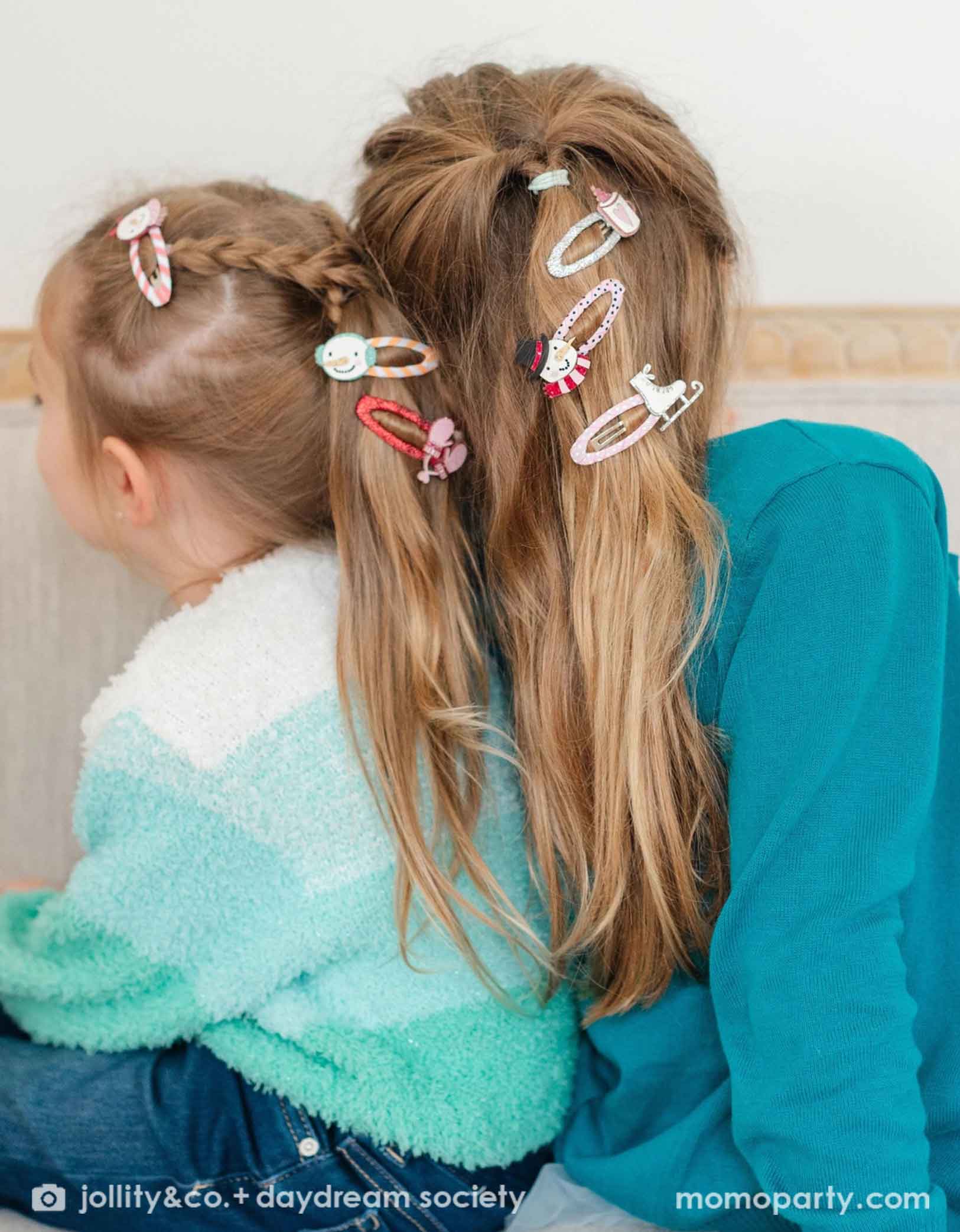 Two girls with colorful holiday winter themed hair clips from Momo Party featuring designs like a snowman, a pair of pink gloves, an ice skate, a cup of hot cocoa in pink in their hair, sitting close together.